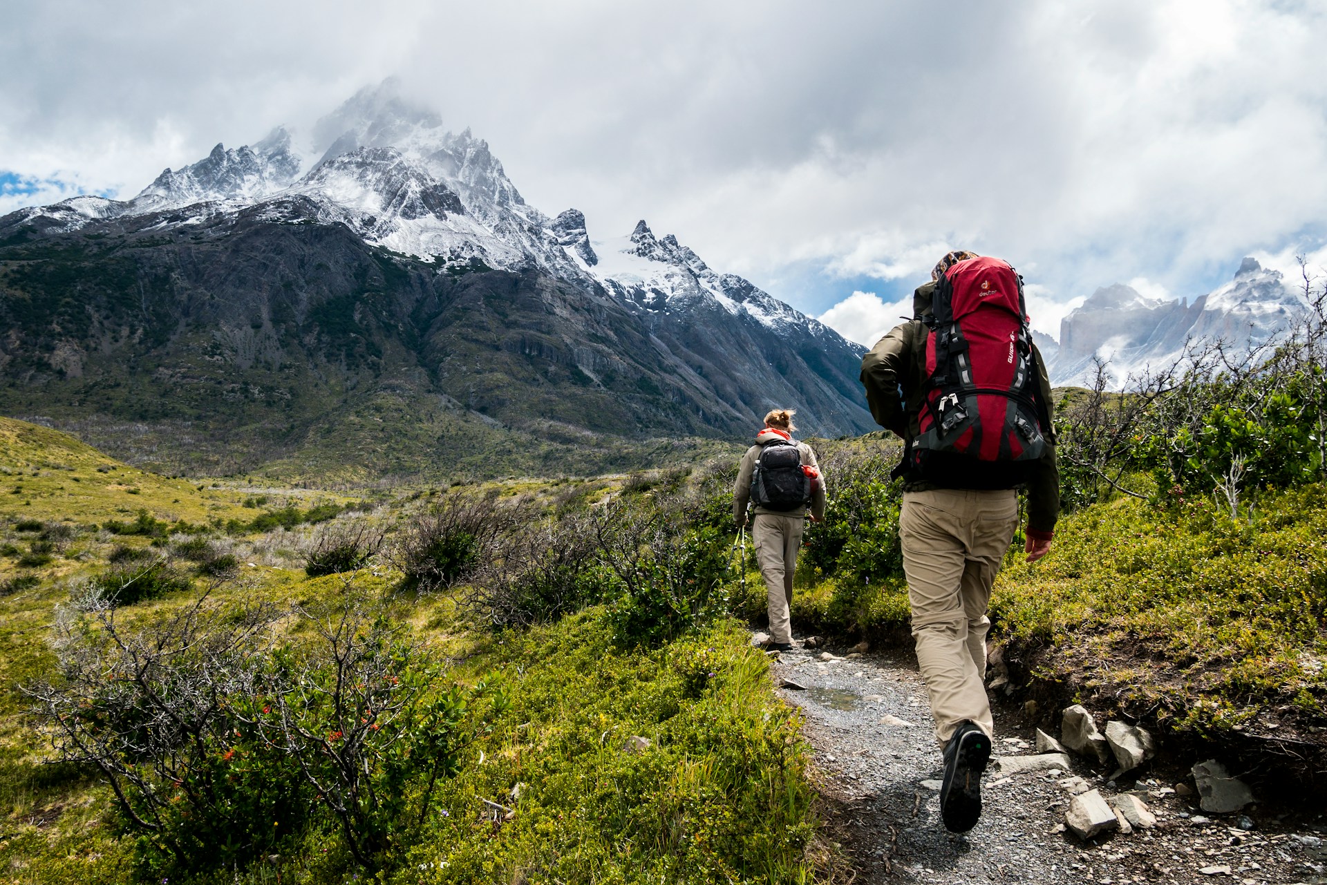 Two people trekking in nature. They are walking in the mountains on a green plain with a view of the snow-capped peaks.